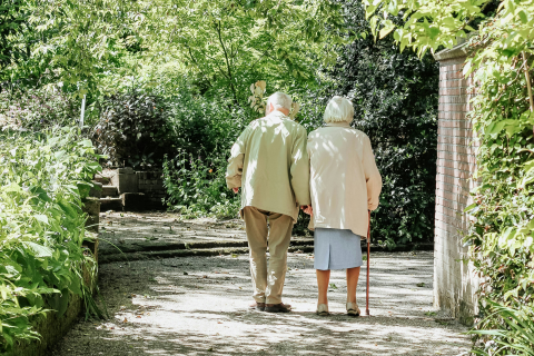 A elderly man and a woman walking together down a lush, green path. The woman wears a skirt and jacket and holds a cane and the man wears slacks and a jacket. 
