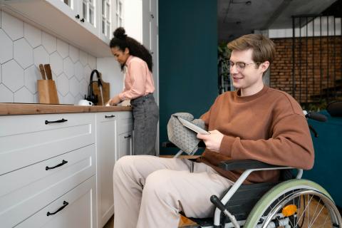 A man with glasses in a wheel chair drying the dishes with a woman standing at the sink in the background