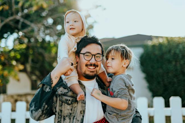 A man with glasses, holding one of his children in one arm and a younger child on his shoulders. 